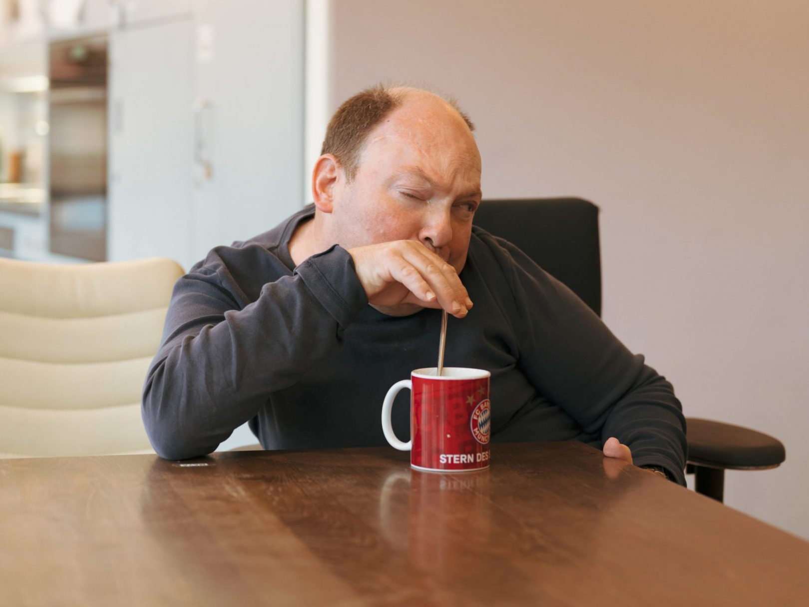 Ein Bewohner aus dem Wohnhaus Wyden sitzt mit zusammengekniffenen Augen am Tisch und trinkt aus einer Tasse.