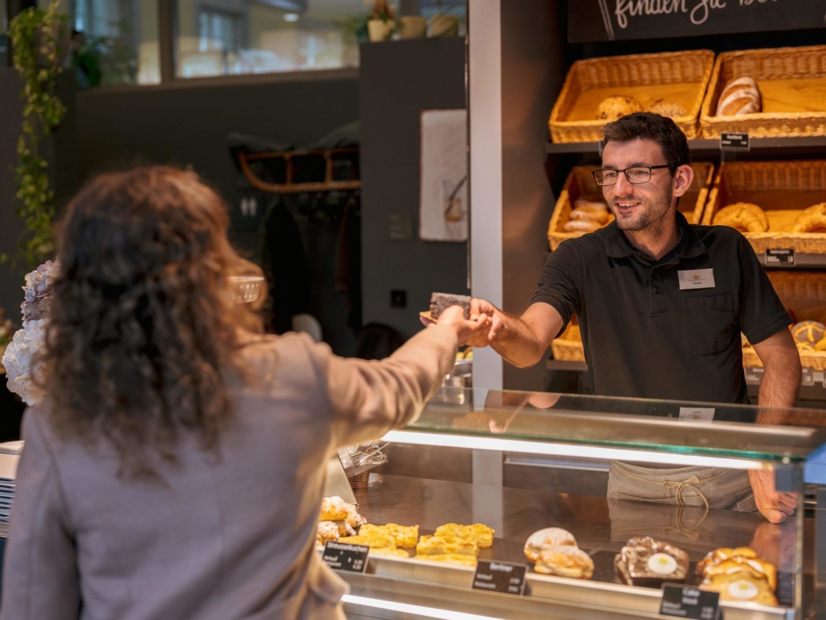 Ein Mann an einer Bäckerei-Theke reicht einer Frau ein Gebäck.