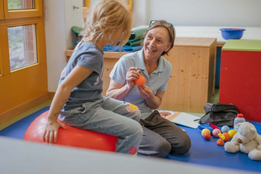 Eine Therapeutin der Brühlgut Stiftung kniet auf einer Matte und arbeitet mit einem Mädchen, das während der Therapiestunde auf einem roten Ball neben ihr sitzt.