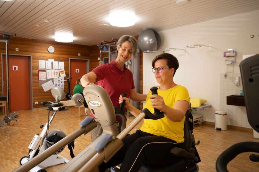 Eine Physiotherapeutin hilft einer Frau, die auf einem Trainingsgerät sitzt.