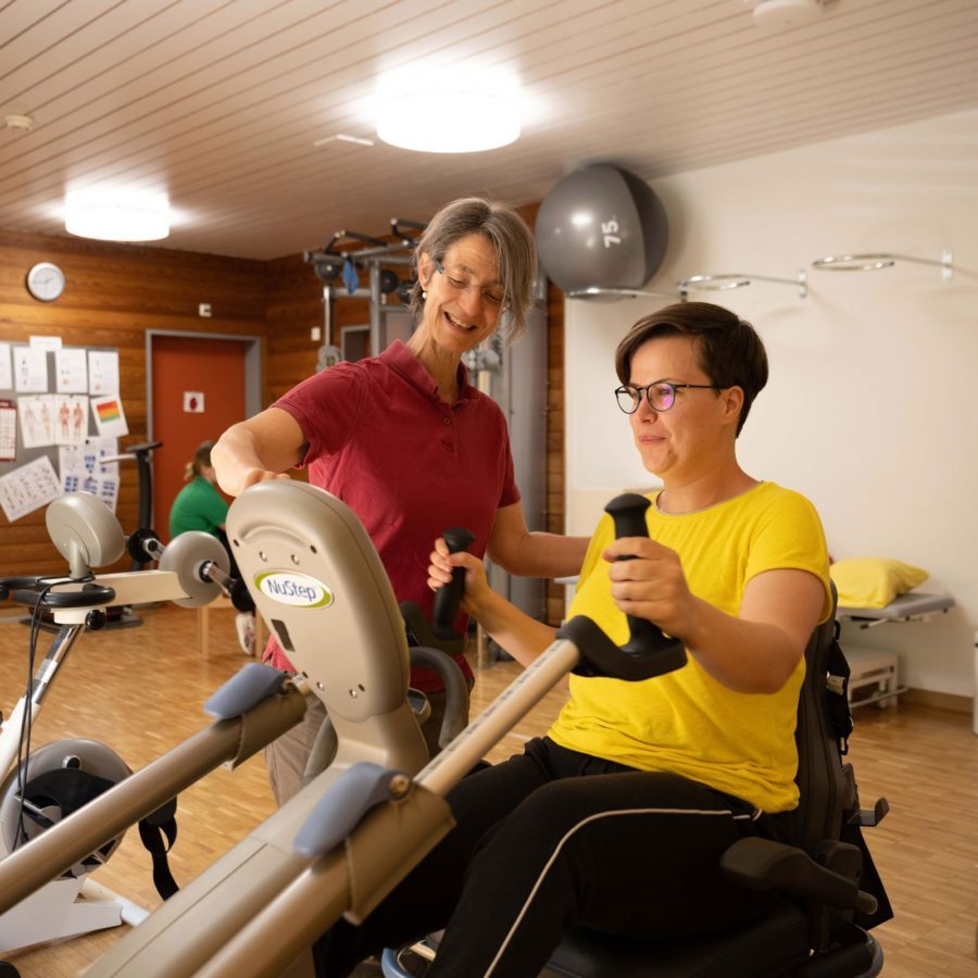 Eine Physiotherapeutin hilft einer Frau, die auf einem Trainingsgerät sitzt.