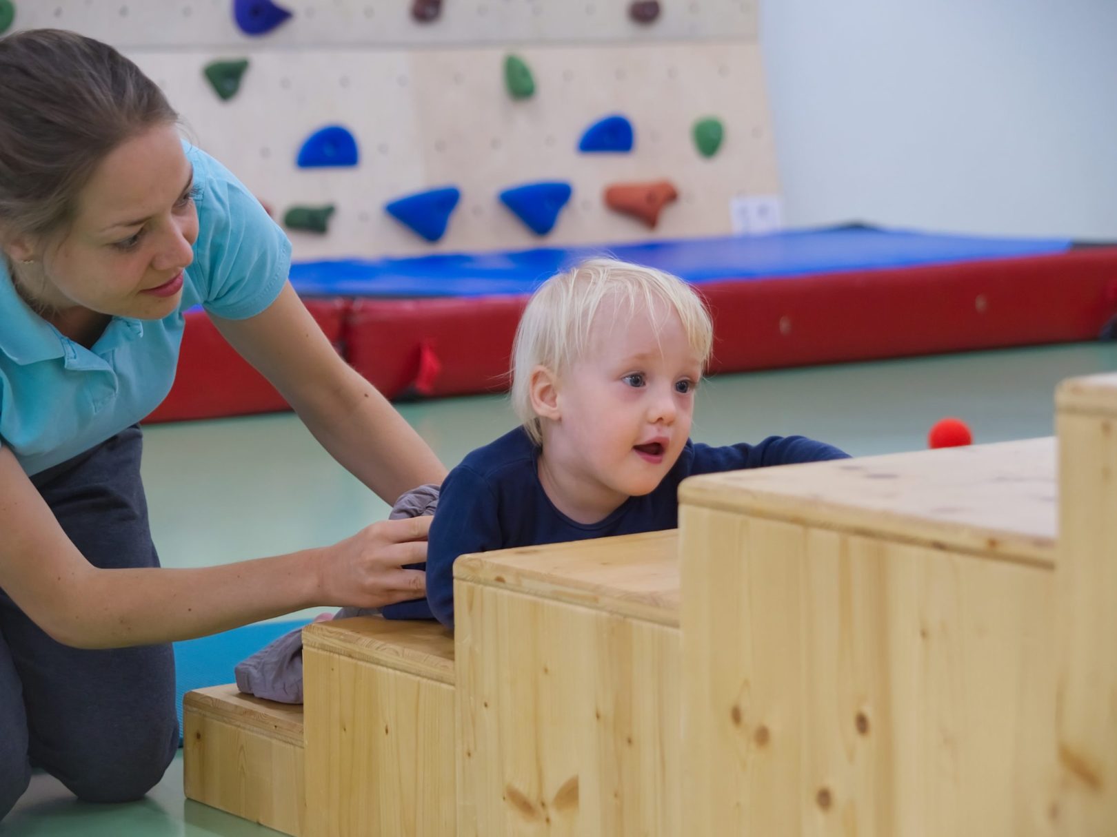 Ein kleiner Junge klettert mit Hilfe einer Physiotherapeutin eine hölzerne Treppe hoch.