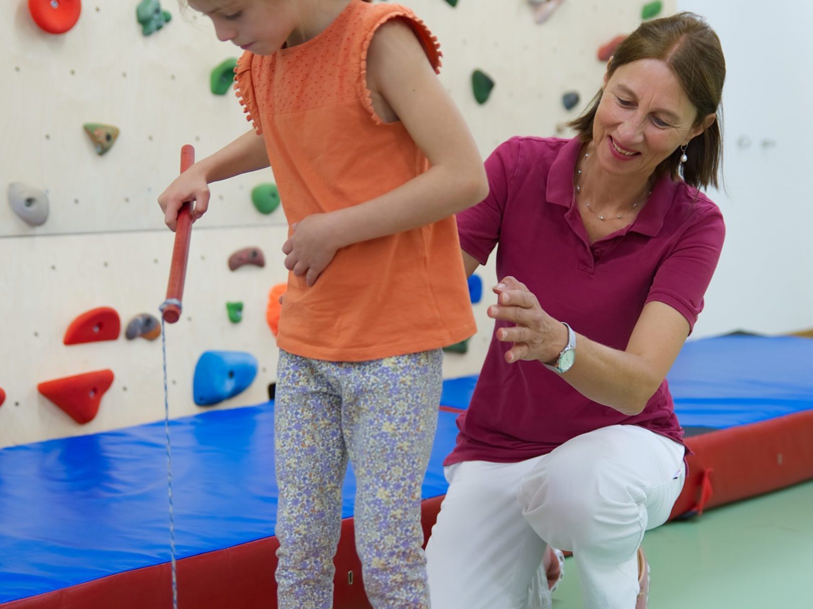 Eine Physiotherapeutin hilft einem stehenden Mädchen bei einem Koordinationsspiel.