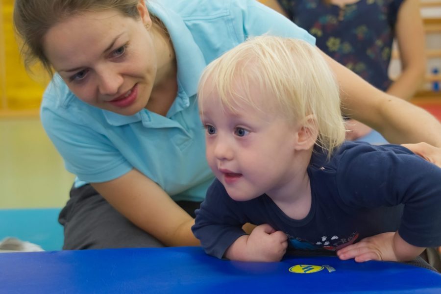 Eine Physiotherapeutin hilft einem blonden kleinen Jungen auf eine Matte zu klettern.