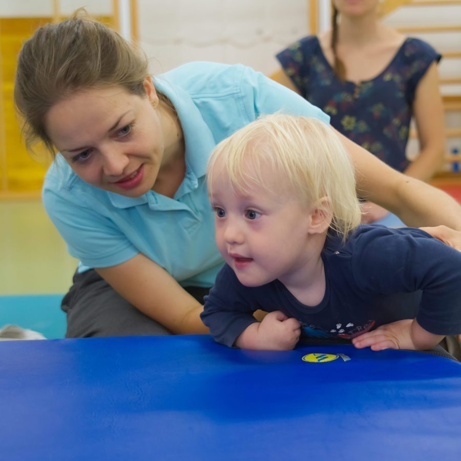 Eine Physiotherapeutin hilft einem blonden kleinen Jungen auf eine Matte zu klettern.