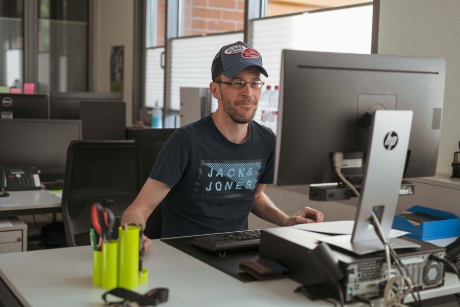 Ein Mann in blauem T-Shirt sitzt an einem Schreibtisch mit einem Computer.