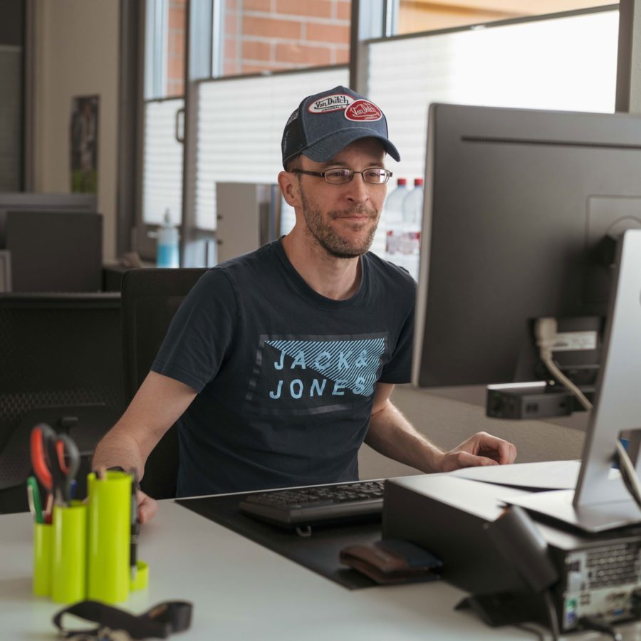 Ein Mann in blauem T-Shirt sitzt an einem Schreibtisch mit einem Computer.