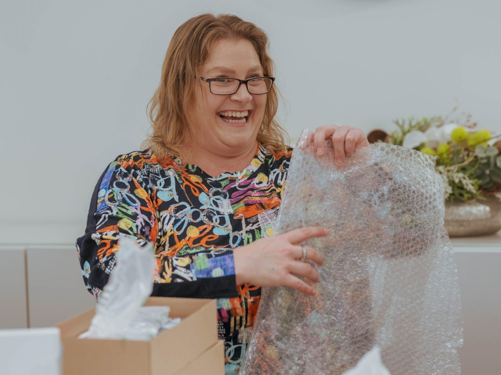 Eine Frau sitzt lächelnd am Tisch und hält Verpackungsmaterial in den Händen.