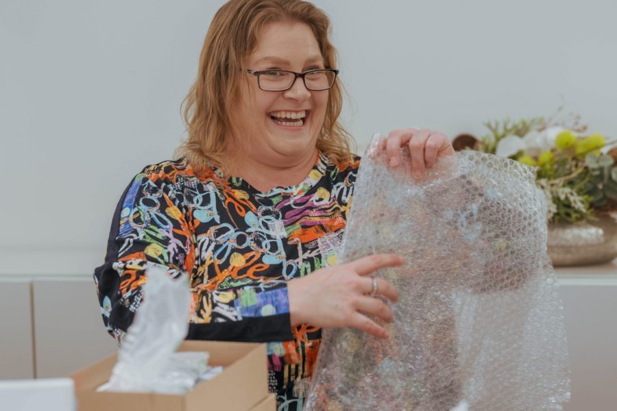 Eine Frau sitzt lächelnd am Tisch und hält Verpackungsmaterial in den Händen.