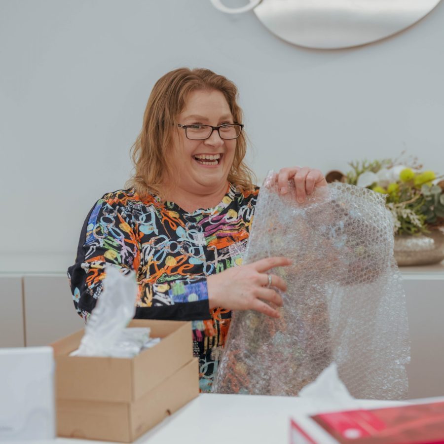 Eine Frau sitzt lächelnd am Tisch und hält Verpackungsmaterial in den Händen.