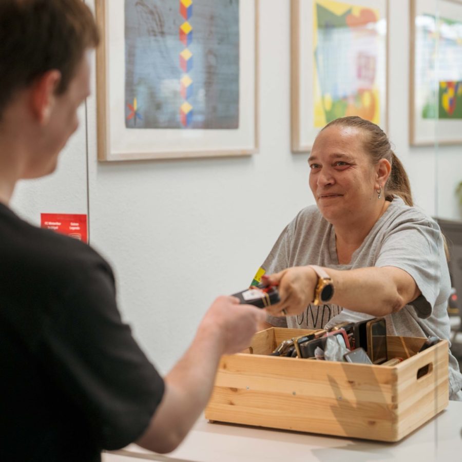 Eine Frau sitzt im Fundbüro und überreicht einem Mann einen Fundgegenstand aus einer Holzbox.