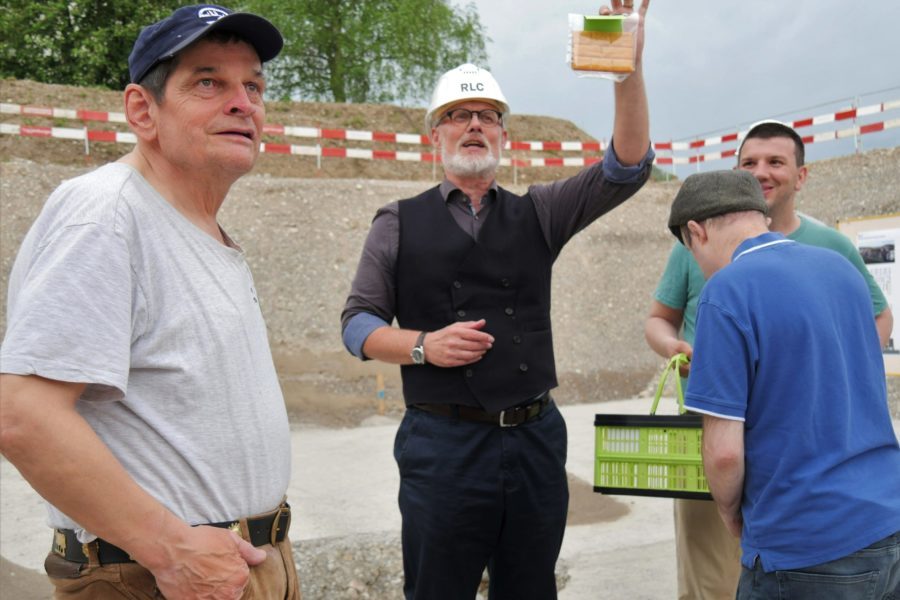 Ein Mann mit Schutzhelm steht in einer Grube und hält den Grundstein der Brühlgut Stiftung in die Höhe. Drei weitere Männer stehen um ihn herum.