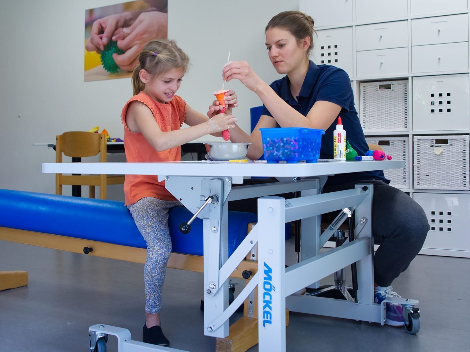 Eine Ergotherapeutin und ein Mädchen sitzen in einem Therapiezimmer und basteln.