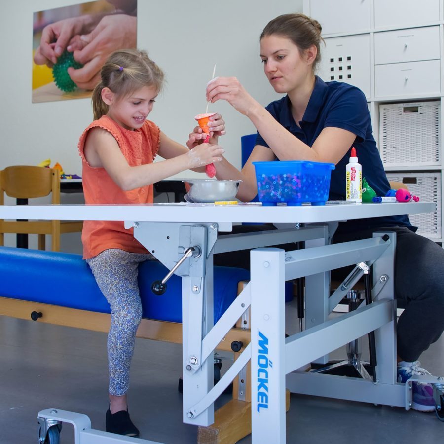 Eine Ergotherapeutin und ein Mädchen sitzen in einem Therapiezimmer und basteln.