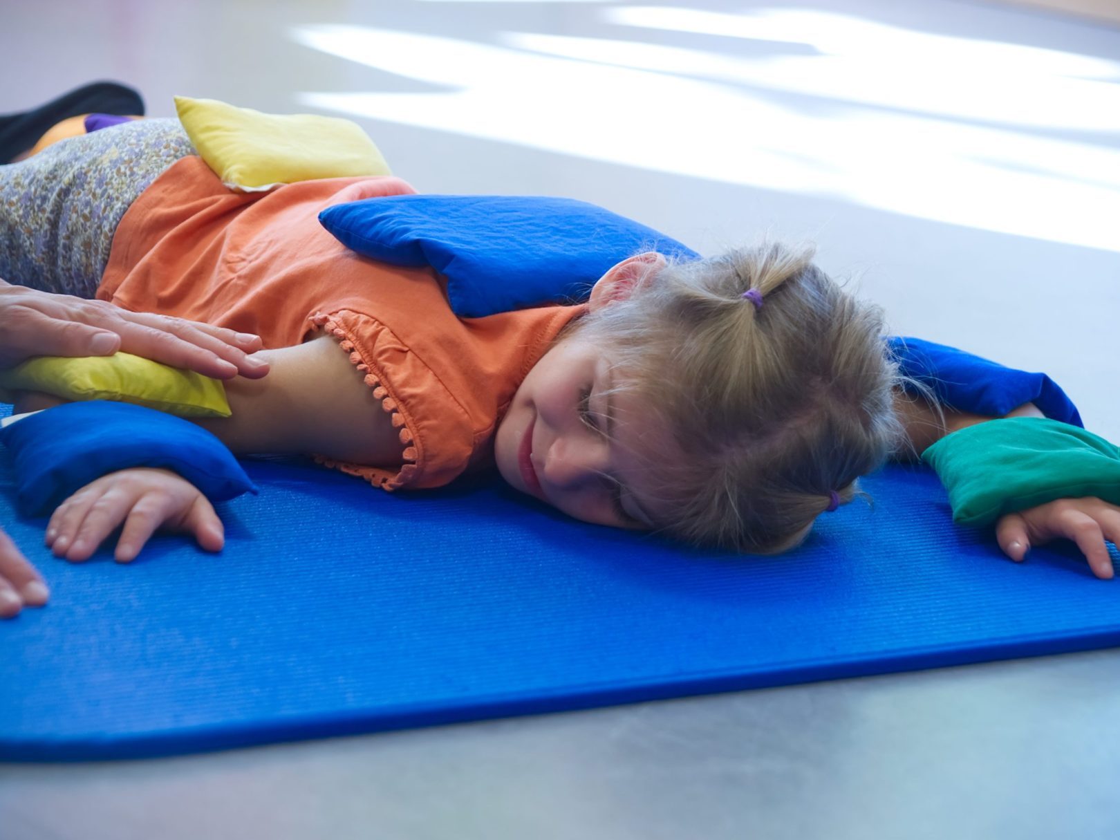 Ein Mädchen liegt auf einer Matte am Boden, während eine Ergotherapeutin kleine Kissen sie legt.