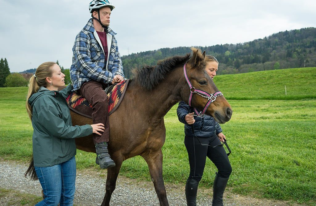 Ein Mann mit Helm reitet während der Hippotherapie auf einem Pferd und wird von zwei jungen Frauen geführt.