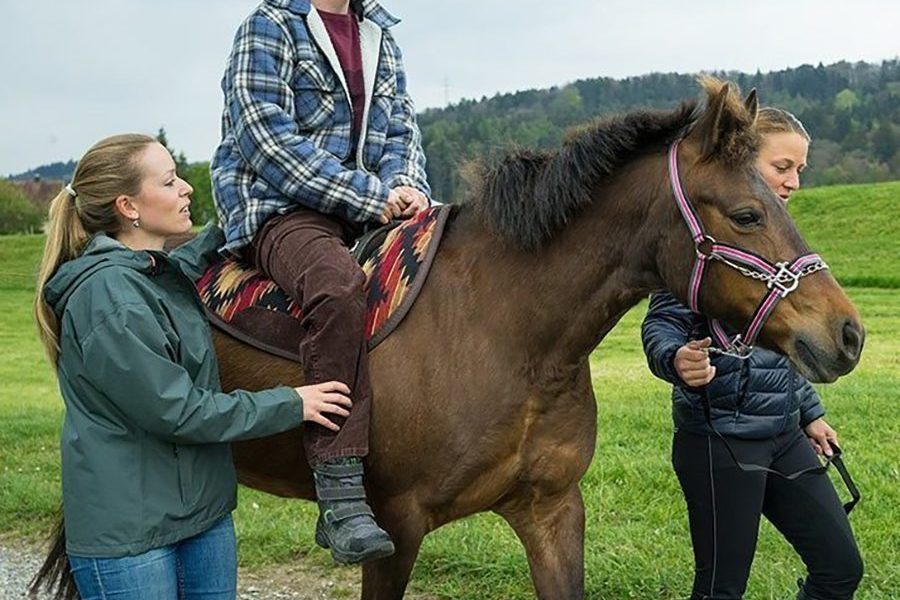 Ein Mann mit Helm reitet während der Hippotherapie auf einem Pferd und wird von zwei jungen Frauen geführt.