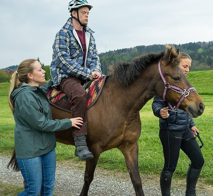 Ein Mann mit Helm reitet während der Hippotherapie auf einem Pferd und wird von zwei jungen Frauen geführt.