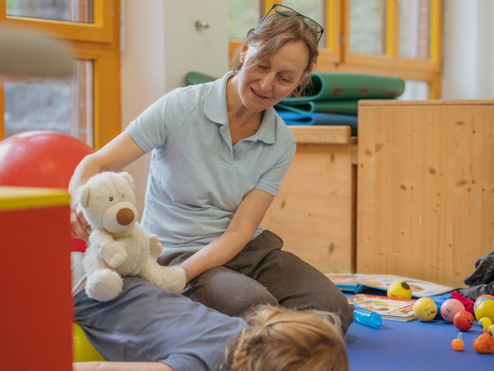Eine Therapeutin setzt ein Plüschtier auf den Rücken eines Kindes, das auf einer Matte liegt.