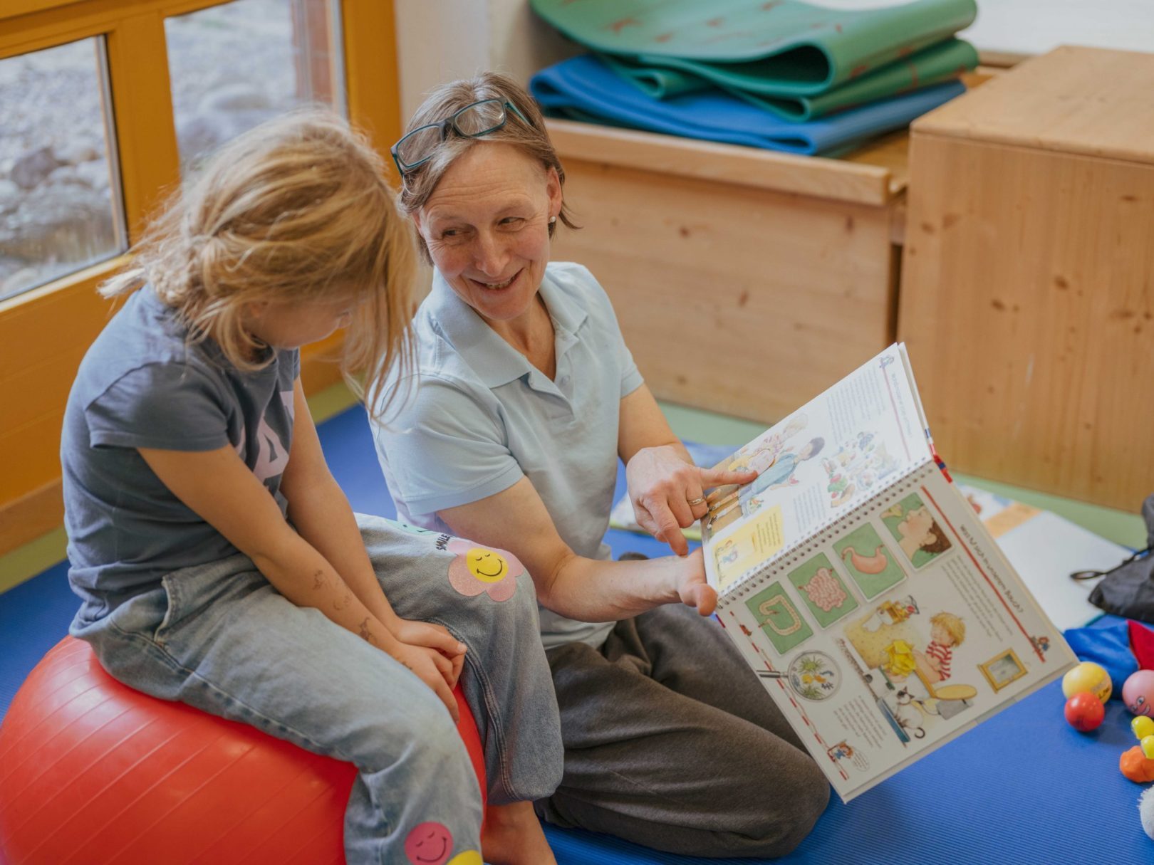 Eine Frau erklärt einem Mädchen in der Urophysiotherapie ein Bilderbuch.