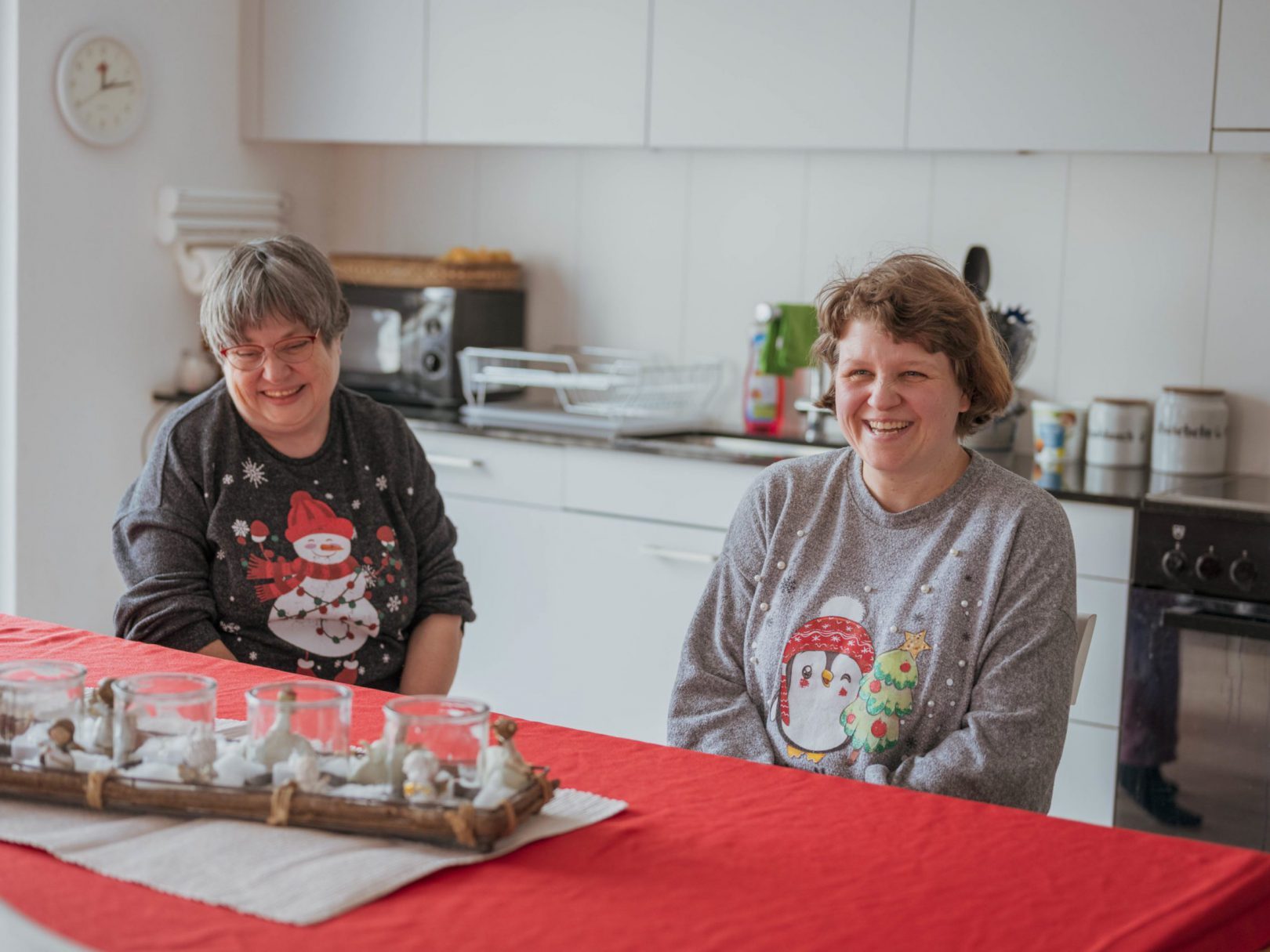 Zwei Bewohnerinnen der Aussenwohngruppe sitzen mit einem Weihnachtspulli am Küchentisch.