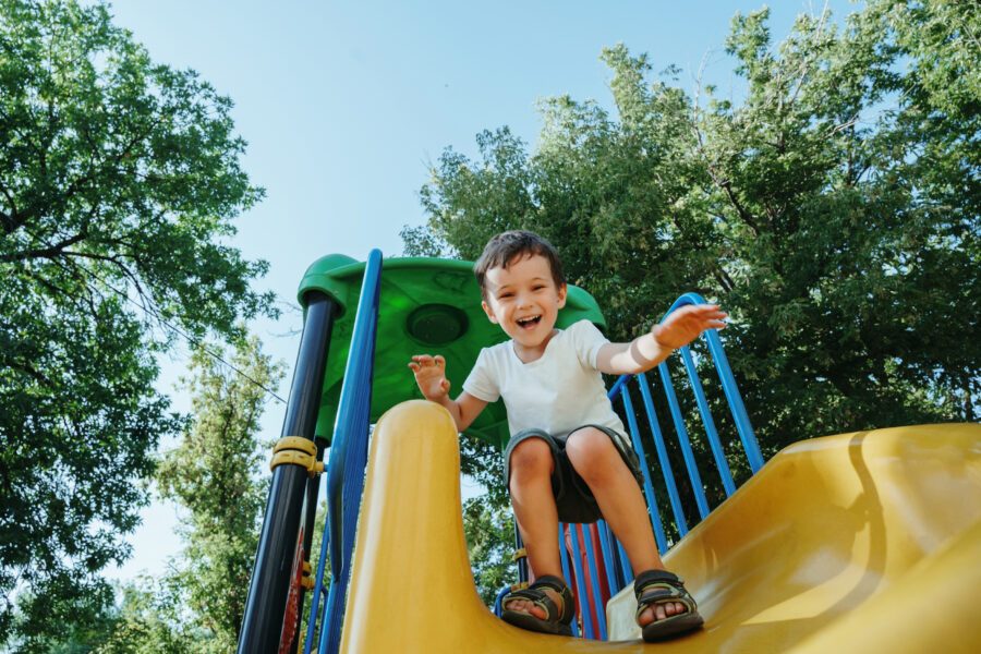 Glückliches Kita-Kind auf einer Rutsche auf dem Spielplatz im Sommer